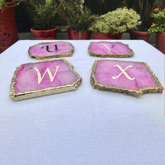 Set of large pink agate drink coasters with gold edges and gold letters U V W X on white surface